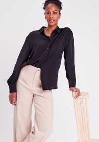 Chemise noire à manches longues, en tissu doux, avec une coupe décontractée, associée à un pantalon large crème et une chaise en bois.