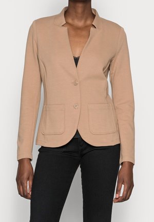 Kobieta ubrana w beżową dopasowaną marynarkę z dwoma przednimi kieszeniami, zapinaną na guziki z przodu, na czarnej bluzce i czarnych spodniach.