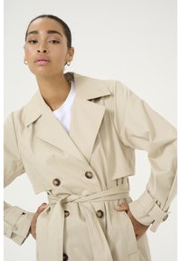 Bežnji trench kaput s dvostrukim kopčanjem sprijeda, velikim ovratnikom i pojasom u struku. Ima teksturiranu tkaninu i smeđe gumbe.