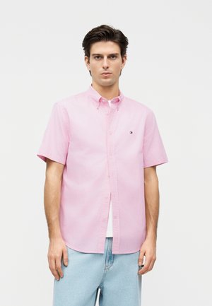 Άνδρας που φορά κοντομάνικο ροζ-άσπρο ριγωτό πουκάμισο με κουμπιά και ανοιχτό μπλε τζιν, στέκεται μπροστά σε μια μονή χρωματική επιφάνεια.