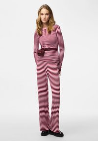 Randig röd och vit jumpsuit med långa ärmar och figursydd midja, kombinerad med ett matchande bälte och en liten väska. Svarta platåskor fullbordar looken.