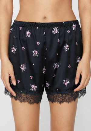 Marineblaue Satin-Shorts mit kleinen rosa und weißen Blumenmustern, mit schwarzem Spitzenbesatz am Saum, getragen von einer Person, die stillsteht.