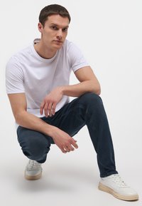 Fehér, rövid ujjú póló, kék farmer és fehér sportcipő. A pólón egy kis logó található a nyaknál; az anyaga sima és laza hatású.