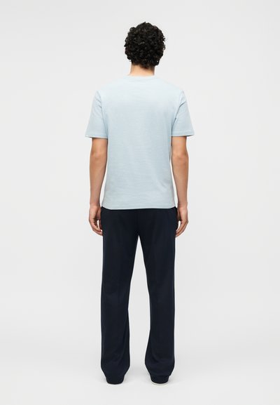 Άνδρας με σγουρά μαλλιά στέκεται με την πλάτη, φοράει ένα ανοιχτό μπλε κοντομάνικο πουκάμισο και φαρδιά σκούρα μπλε παντελόνια με φόντο μονόχρωμο.