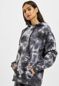 Felpa grigia tie-dye presenta un cappuccio con coulisse, tasca frontale e motivo logo a scritta. Realizzata in tessuto morbido e texture.