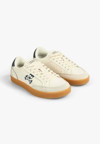 Baskets en cuir beige avec une semelle en caoutchouc gomme, bout arrondi et un logo de tête de mort avec des os croisés sur le côté. Présente des lacets blancs et des accents texturés.