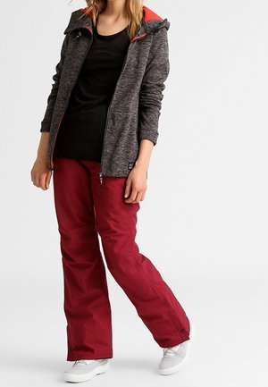 Gray zip-up jacket with red accents, black shirt, and burgundy pants. Model wears white sneakers. Textured fabric, casual design.