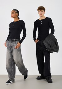 Dva posameznika predstavljata črne long-sleeve henley majice. Eden nosi svetlo sive ohlapne kavbojke in črn pas; drugi ima temne ohlapne kavbojke in jakno.