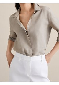 Chemise en lin gris clair à boutons, coupe décontractée et manches retroussées, associée à un pantalon blanc sur mesure avec un détail ceinturé.