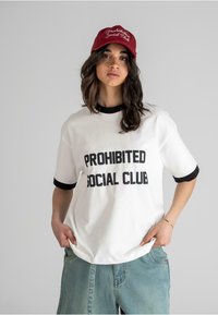 Biela bavlnená tričko s čiernym textom "PROHIBITED SOCIAL CLUB", s čiernym rebrovaným golierom a rukávmi. V kombinácii s vyblednutými modrými džínsovými šortkami.