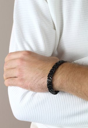 Schwarzes geflochtenes Armband aus gummiartigem Material, mit ineinander verflochtenen Gliedern und einer glatten Oberfläche, getragen am Handgelenk vor einem weißen Stoffhintergrund.