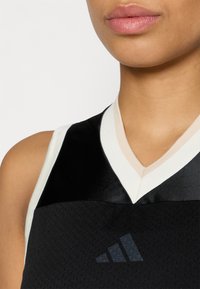 Maglia sportiva nera con scollo a V, caratterizzata da un tessuto strutturato e un bordo bianco. Logo Adidas in colore scuro sul davanti.