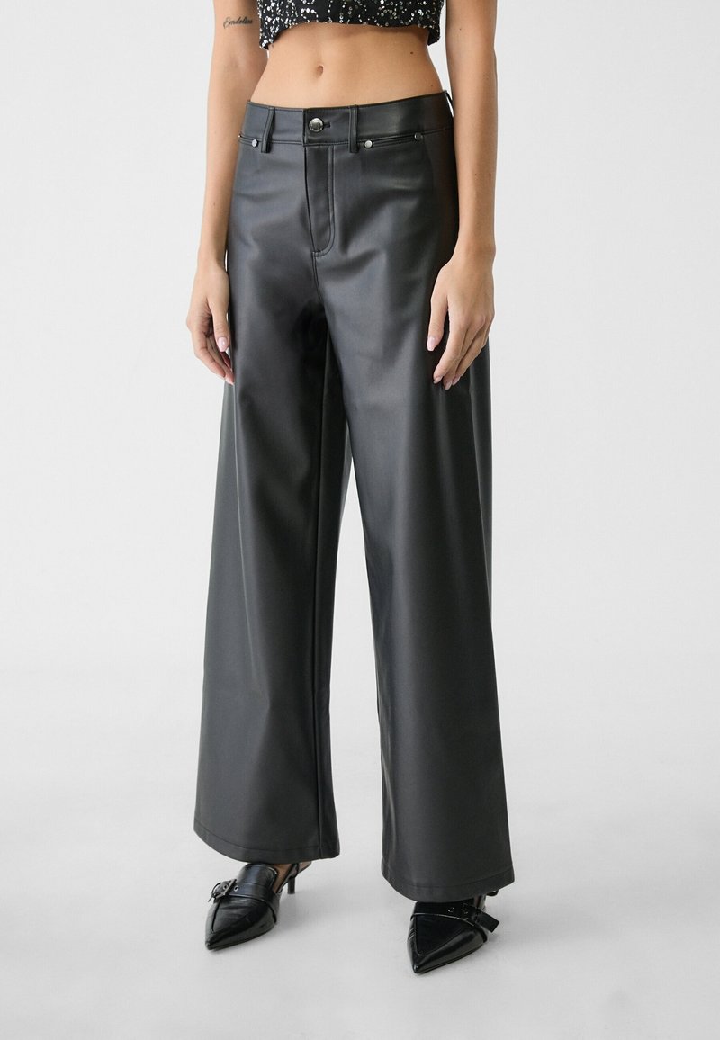 Femme portant un pantalon en cuir noir taille haute à jambes larges, des talons noirs pointus, et un top court à motifs, debout devant un fond uni.
