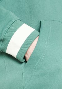 Zelená mikina s bílými pruhovanými manžetami a boční kapsou, vykazující hladkou texturu a pečlivé švy.