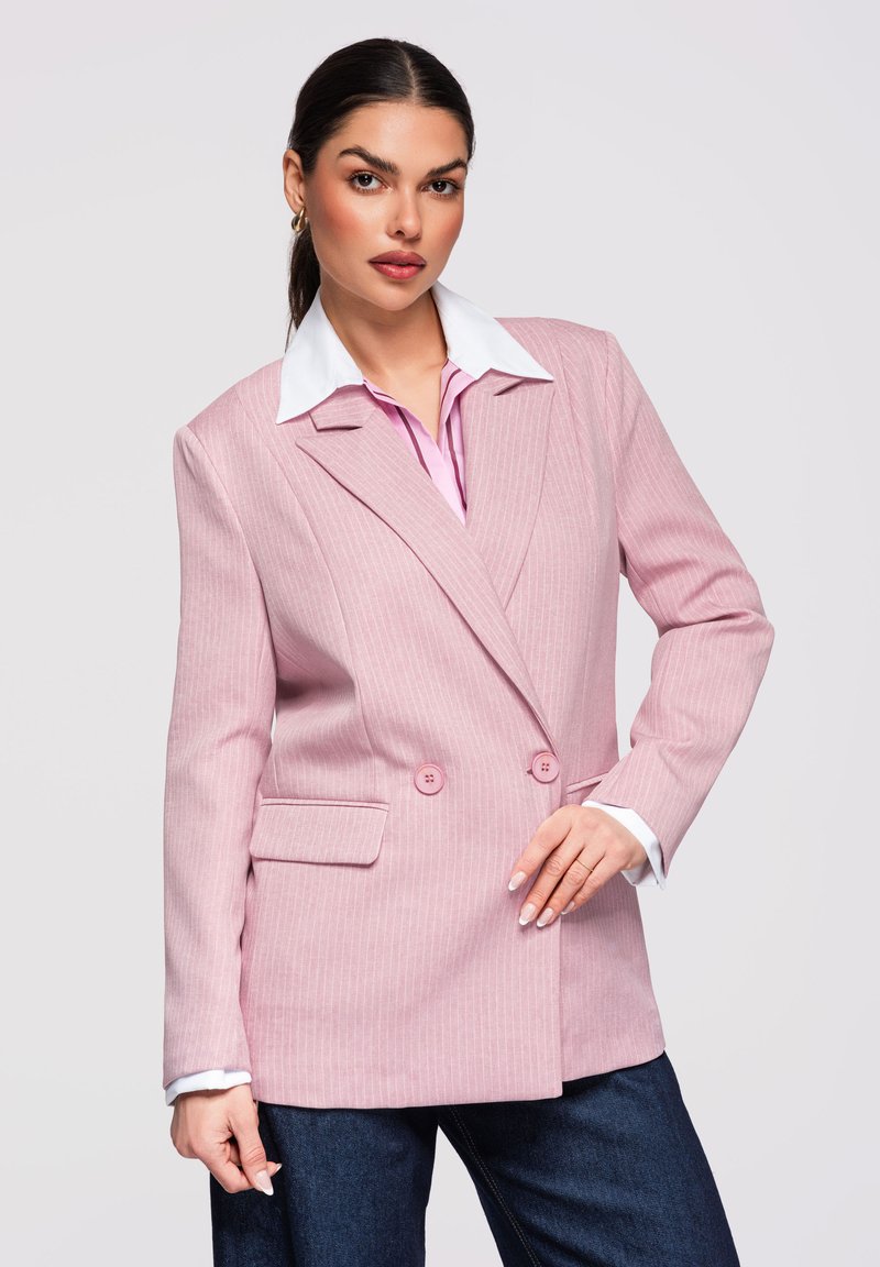 Femme aux cheveux foncés portant un blazer croisé à fines rayures rose sur une chemise à col blanc et un jean foncé, posant devant un fond uni.