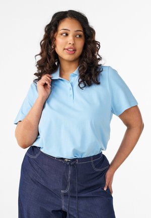 Žena s kučeravými vlasmi, oblečená v svetlomodrej polo košeli a tmavomodrých nohaviciach s vysokým pásom, pozerá sa do strany s jednou rukou na bedre.