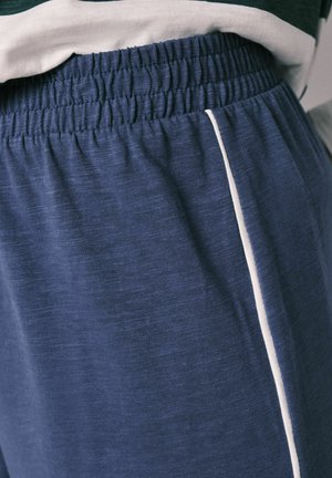 Ceinture élastique sur un pantalon bleu marine avec un liseré blanc sur le côté, porté avec un haut rayé blanc et bleu marine rentré à l'intérieur.