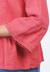 Korallefarbene Leinenbluse mit Rundhalsausschnitt und Dreiviertelärmeln, die am Bündchen mit einem zarten Spitzenrand und am Saum mit Gathered-Details versehen ist.