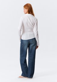 Camicia bianca a maniche lunghe con tessuto strutturato, abbinata a jeans di denim blu larghi. L'outfit ha una silhouette casual e rilassata.