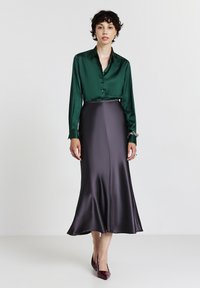 Blusa in satin verde con chiusura a bottoni, abbinata a una gonna in satin viola scuro fluente. Le calzature sono dei tacchi alti borgogna scuri.