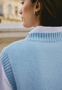 Lichtblauwe gebreide vest met ribbelachtige halslijn, gecombineerd met een witte blouse met kraag. Zachte textuur, casual ontwerp en subtiele laagdetails.