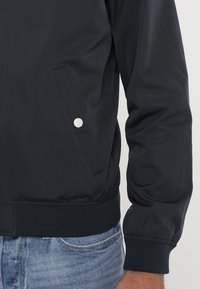 Chaqueta bomber de color navy con una textura suave, puños acanalados y un bolsillo lateral con detalle de botón a presión plateado. Usada sobre pantalones de mezclilla.