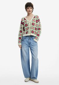 Virkat kardi med färgglada blommotiv i rosa och grönt, korta ärmar och V-ringning, kombinerat med lösa blå jeans och beiga skor.