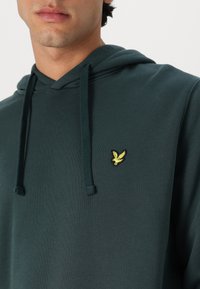 Zelená mikina s kapucňou vyrobená z jemnej tkaniny, s kapucňou na sťahovanie a malým žltým vyšitým logom vtáka na hrudi.