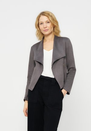 Ženska s svetlo rjavimi lasmi, oblečena v sivo odprto jakno, belo bluzo in črne hlače z visokim pasom, stoji na enostavni bež ozadju.