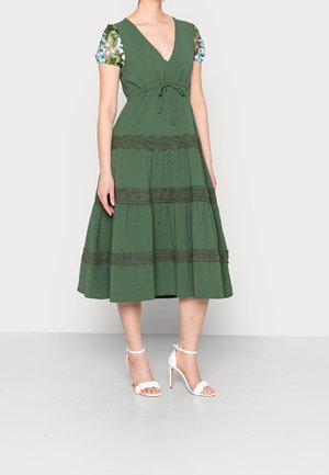 Femme portant une robe midi verte avec des bandes en dentelle et des manches courtes fleuries, associée à des sandales blanches à talons hauts.
