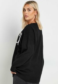 Svart stickad tröja med avslappnad passform, ribbad nederkant och ärmslut, med vit text på framsidan. Mjuk textur, rundad halsringning.