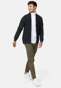 Mørkegrå strikket cardigan med åbent frontdesign, ribbet tekstur, båret over en hvid turtleneck, parret med olivengrønne bukser og hvide sneakers.