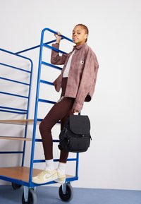 Brown leggings, light gray top, and a mauve button-up jacket. Black backpack with a flap. Yellow Nike sneakers with white socks. Blue storage rack.