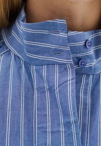 Blå randig skjorta med hög krage. Tyget är lätt och texturerat. Har två lila knappar och en knappslå med knapphål.