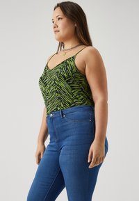 Camiseta sin mangas de patrón verde y negro con tirantes finos, combinada con unos vaqueros de mezclilla azul con cierre de cremallera y bolsillos frontales.
