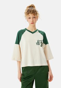 Cremefarvet og grøn kortærmet V-hals T-shirt med raglanærmer, der har "New York 91" trykt i grøn og en afslappet pasform.