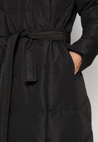 Svart quiltad kappa med bälte knutet vid midjan, ena handen i sidofickan, närbild som visar tygtextur och sömnadsdetaljer.