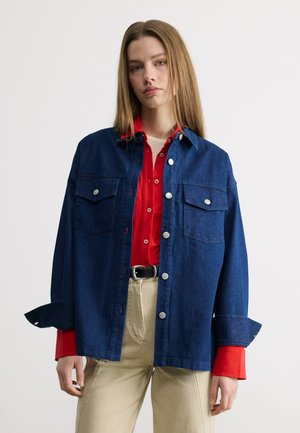 Fiatal nő egyenes hosszú hajjal, sötétkék farmerdzsekit visel, piros gombos inggel és bézs nadrággal, fekete övvel.