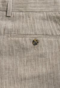 Poche d'un pantalon beige à fines rayures, avec un bouton marron foncé unique et une boucle de ceinture en tissu au-dessus.