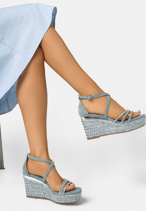 Woman wearing light blue braided wedge sandals with ankle straps and rhinestone embellishments, crossed leg, light blue skirt visible.