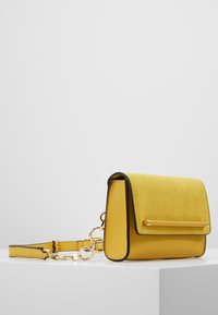 Borsa in suede gialla con un design strutturato, rifiniture nere e un accento dorato sulla patta. Presenta una tracolla removibile.