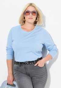 Světle modrá dlouhá košile, černý cvočkovaný opasek, šedé džíny. Model má na sobě velké fialové sluneční brýle a drží šedou tašku.