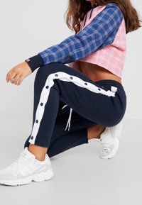 Felpa cropped a scacchi rosa e blu, abbinata a pantaloni jogger blu notte con dettagli a bottoni bianchi lungo i lati. Sneakers sportive bianche.