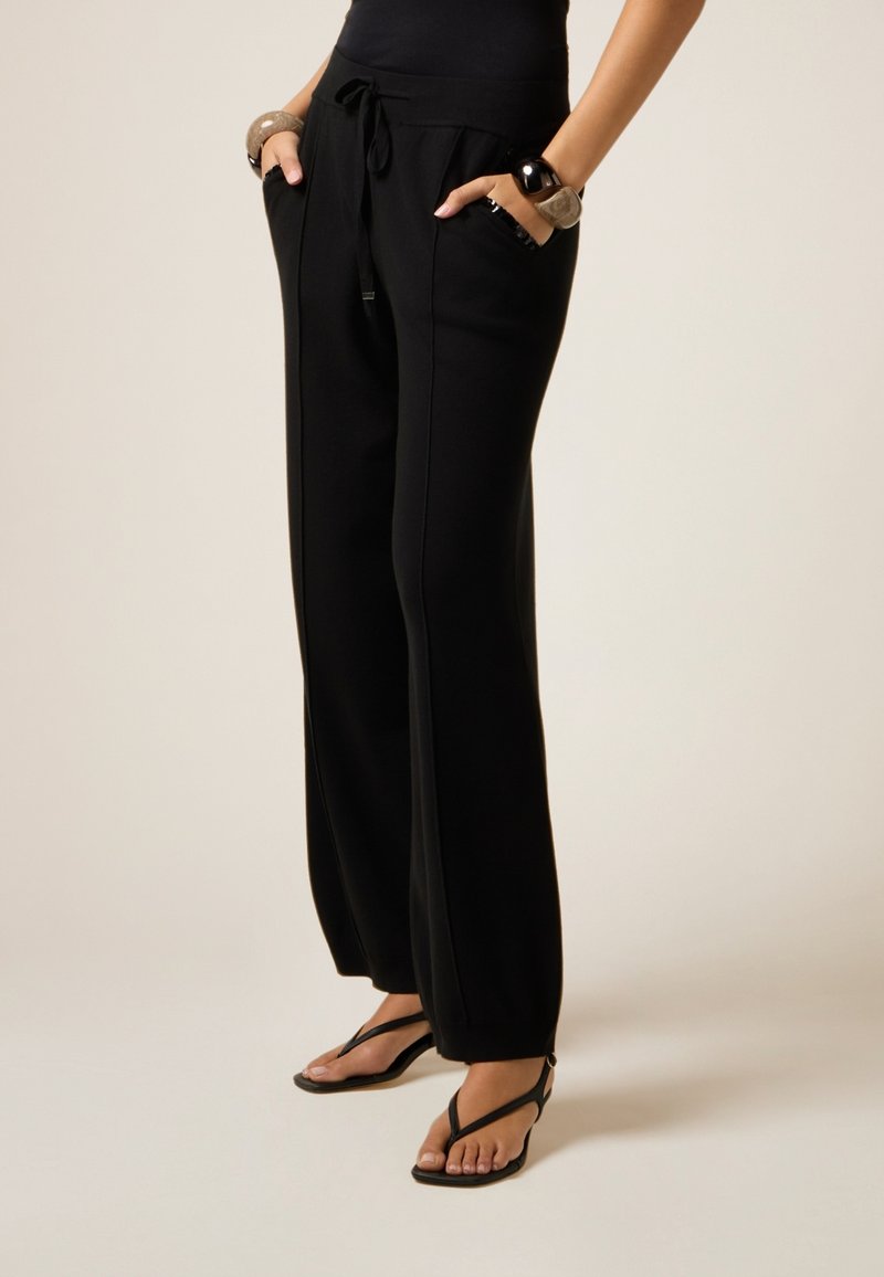 Zwarte sweatpants met een trekkoord in de taille, zijzakken en een losse pasvorm. Gecombineerd met zwarte slippers op een neutrale achtergrond.