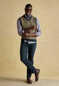 Kootud vest, millel on houndstooth muster maapinna toonides, üle triibulise särk, koos tumedate teksade ja pruunide nokatsitega.