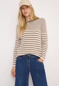 Pull à manches longues rayé beige et marron, avec une texture côtelée. Associé à un jean large bleu foncé. Détails avec une casquette.