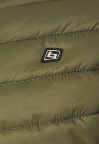 Gros plan sur un tissu matelassé vert olive avec un petit patch logo rectangulaire noir et blanc cousu dessus.