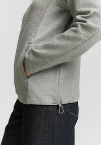 Grijze textuurjacket met een gladde gebreide body, geweven accenten op de schouders, zijzakken en een lusdetail aan de zoom.