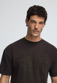 Brun bomullströja med korta ärmar, som har en tonad "POLO CLUB"-tryck fram, rund halsringning och en avslappnad passform.