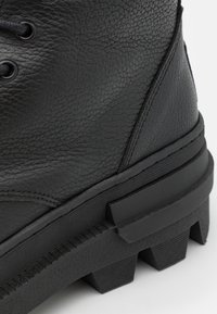 Gros plan sur une botte en cuir noir texturé avec une semelle crantée épaisse et des coutures détaillées sur fond blanc.